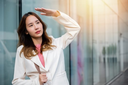 Portrait of confidence young businesswoman standing outside office building in city raise her hand to shade sun UV. Happy woman wearing white suit jacket with brown bag at sunlight outdoors. sun block