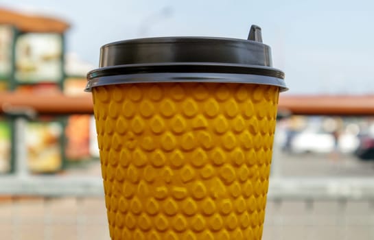 One yellow paper glass of tea or coffee - takeaway food on a table in a cafe. Nobody. A disposable glass with a hot drink stands against the backdrop of a summer cafe