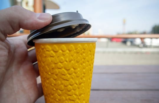 One yellow paper glass of tea or coffee - takeaway food on a table in a coffee shop. A hand covers a disposable glass with a lid against the backdrop of a summer cafe