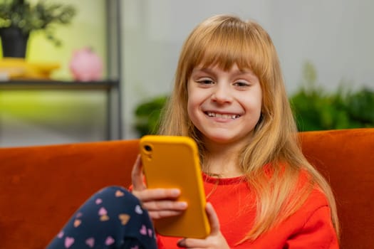 Young child girl texting share messages content on smartphone social media applications online watching relax movie. Smiling teenager little kid uses mobile phone at home in living room sits on sofa
