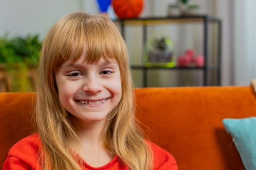 Close-up portrait of happy smiling Caucasian preteen school girl 8 years old. Young lovely blonde little child kid looking at camera at home play living room apartment sitting on couch. Copy-space