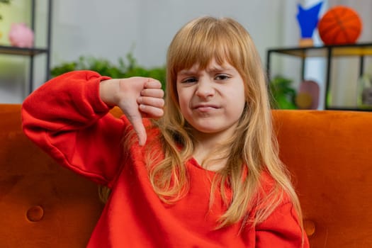 Dislike. Upset child girl showing thumbs down sign gesture, expressing discontent, disapproval, dissatisfied bad work at modern home apartment indoors. Displeased teenager kid in play room on sofa