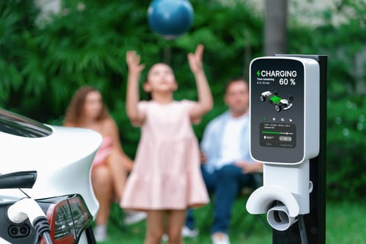 Focus electric vehicle recharge from home charging station on blur background of happy and playful family playing together. EV car using alternative and sustainable energy for better future.Synchronos