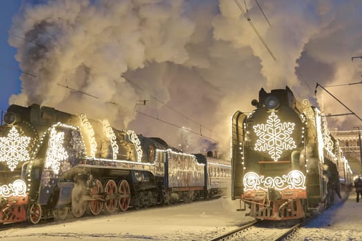 Russia, Peterhof, 07 January 2024: Two Santa Claus Trains leave the magically decorated building of the New Peterhof station, huge steam clubs, many people, incredible lighting. High quality photo