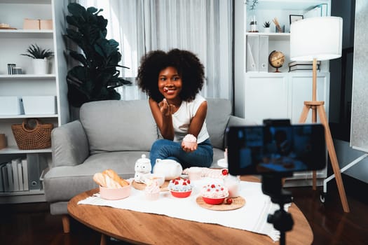 Beautiful young African blogger presenting piece of cupcake in concept special cuisine with video record on smartphone. Content creating of social media with favorite sweets bakery dish. Tastemaker.