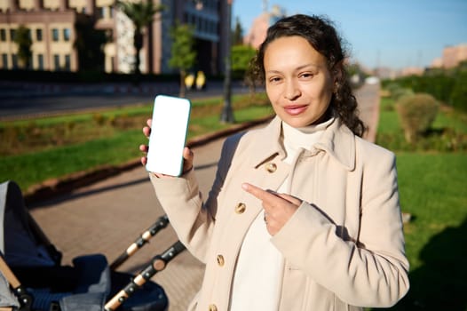 Happy multi ethnic smiling woman standing on the city square, looking at camera and pointing her finger at white blank digital screen on her smartphone, copy advertising space for mobile app . Mockup.