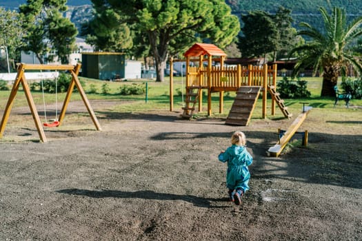 Little girl goes to a wooden playground in a green park. Back view. High quality photo