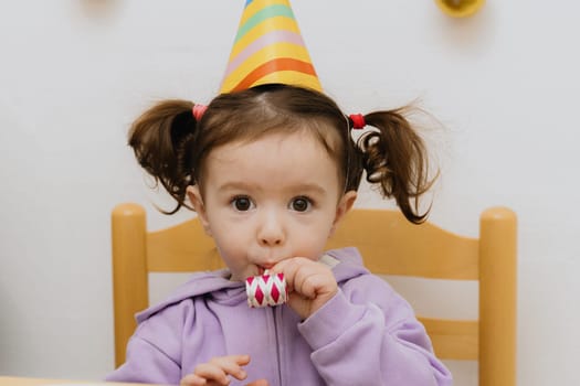 One beautiful Caucasian girl with a cone hat on her head sits at a festive table and blows into a paper tube, looking at the camera and celebrating her birthday, close-up side view.