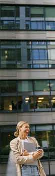 Sales manager standing with laptop and coffee on office terrace during break time and looks away