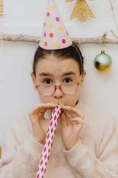 One beautiful Caucasian brunette girl with glasses and a pale pink cone hat on her head sits on a chair at a festive table and blows hard into a paper tube, looking cheerfully at the camera and celebrating her birthday, close-up side view.