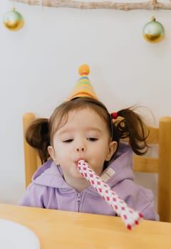 One beautiful Caucasian girl in a conical hat and two ponytails on her head sits at a festive table and blows strongly into a paper pipe, looks to the side, celebrates her birthday, close-up side view.