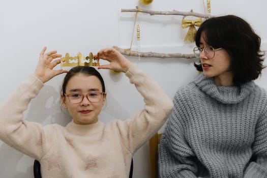 One beautiful Caucasian girl with glasses solemnly puts a golden paper crown on her head, after eating a royal galette, sitting at the table in the kitchen with her family, close-up side view.