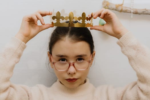 One beautiful Caucasian girl with glasses solemnly puts a golden paper crown on her head, after eating a royal galette, sitting at the table in the kitchen, close-up view from above.