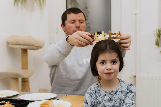 One young handsome Caucasian man puts a golden paper crown on his little girl daughter after eating a royal galette and finding a gift, while sitting at the table in the kitchen, close-up side view.