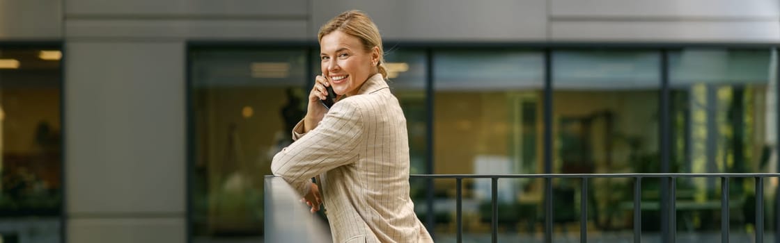 Smiling female manager talking phone standing on modern office building background