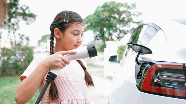 Happy little young girl learn about eco-friendly and energy sustainability as she recharge electric vehicle from home EV charging station. EV car and sustainable future generation concept. Synchronos