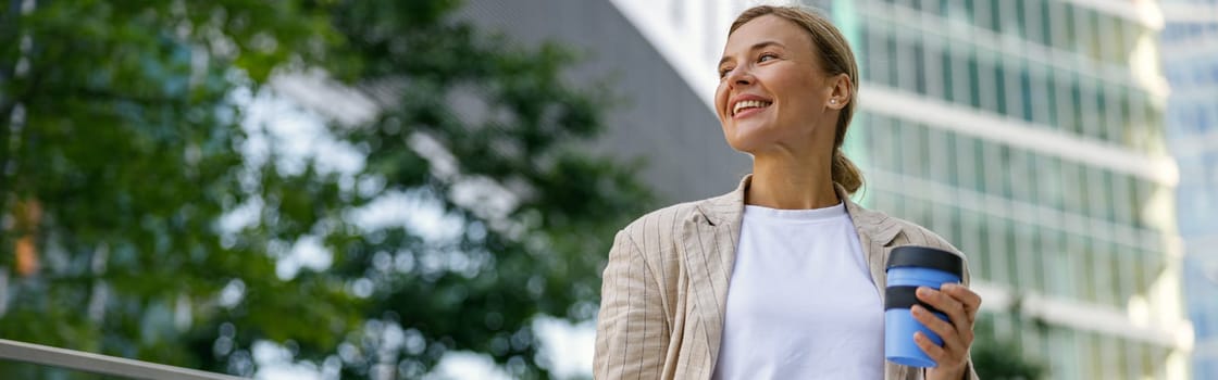Smiling business woman drinking coffee during break time near office building and looks away