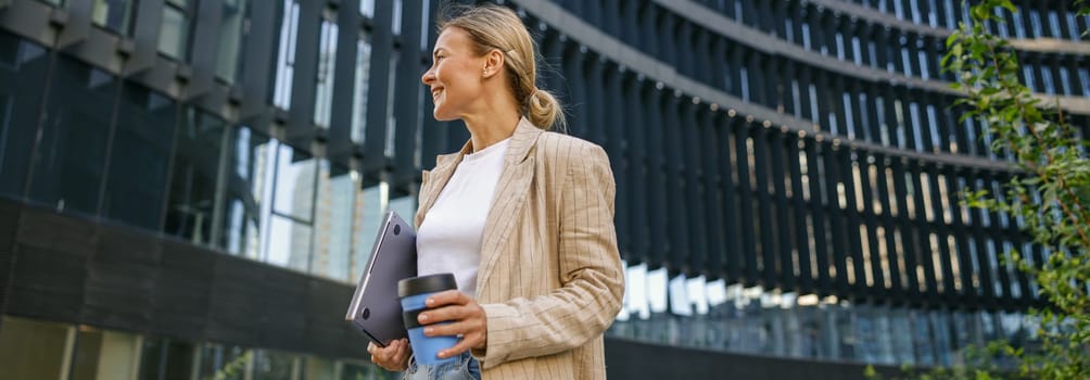 Positive female manager with laptop and take away coffee during break time near office building