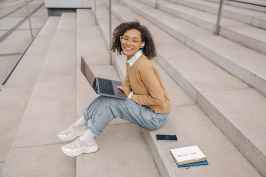 Pretty female entrepreneur working on laptop sitting outside on modern building background