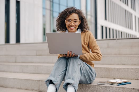 Pretty female entrepreneur working on laptop sitting outside on modern building background