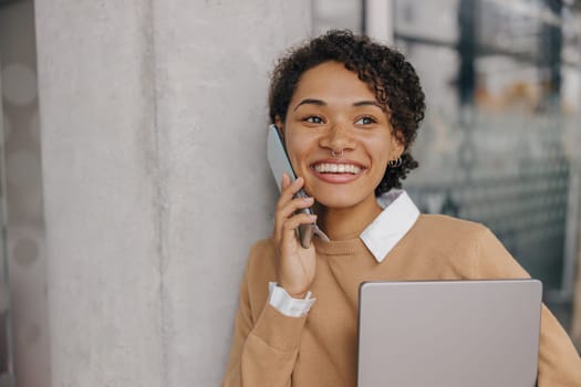 Stylish business woman is talking phone while standing with laptop in modern office