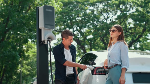 EV car recharging electricity for battery by lovey couple traveling during their road trip travel by eco friendly electric vehicle in national park and greenery forest on holiday. Exalt