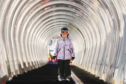 A girl on skis stands on a carpet ski lift in a tunel