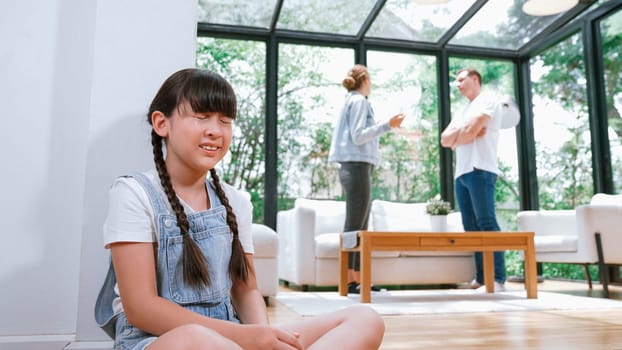 Stressed and unhappy young girl huddle in corner, cover her ears blocking sound of her parent arguing in background. Domestic violence at home and traumatic childhood develop to depression. Synchronos