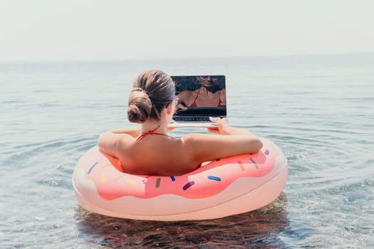 Woman freelancer works on laptop swimming in sea on pink inflatable ring. Pretty lady typing on computer while floating in the sea on inflatable donut at sunset. Freelance, remote work on vacation