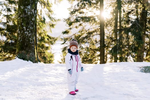 Little girl in a ski suit stands sideways on the edge of a snowy forest and looks away. High quality photo