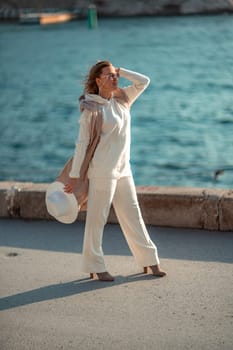 Happy blonde woman in a white suit and hat posing at the camera against the backdrop of the sea.