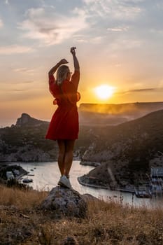 Happy woman standing with her back on the sunset in nature in summer with open hands posing with mountains on sunset, silhouette. Woman in the mountains red dress, eco friendly, summer landscape active rest.