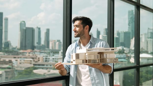 Skilled smiling caucasian engineer turns house model left and right to check mistake point. Professional manager inspect at architectural model while standing near window. Design concept. Tracery