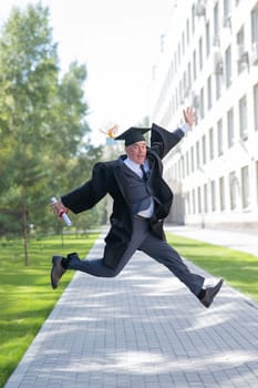 Old happy man in graduation gown jumping outdoors and holding diploma. Vertical