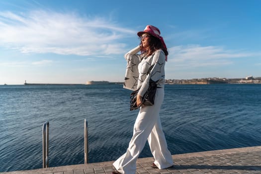 Stylish seashore woman. Fashionable woman in a hat, white trousers and a light sweater with a black pattern on the background of the sea