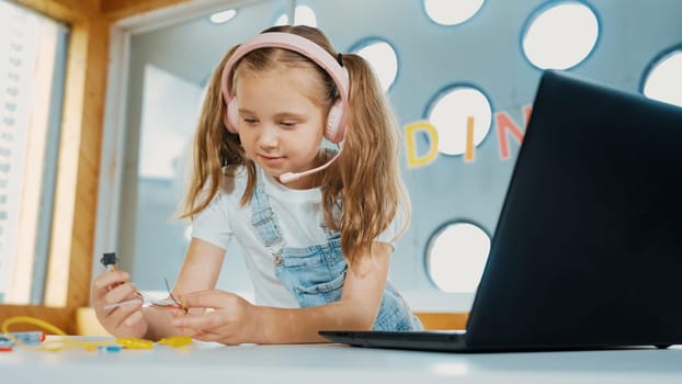 Pretty girl wearing headphone while study electronic equipment. Caucasian child doing science experiment while laptop, screwdriver and wires placed near on table. Smart online classroom. Erudition.