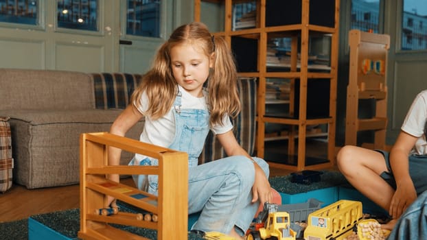 Caucasian girl sits at sand box while placed car model at slope toy. Diverse children playing plastic model while sitting at school in break time at play room. Creative activity concept. Erudition.