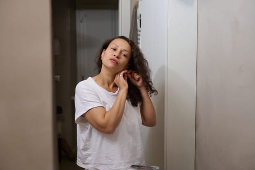 Beautiful curly haired brunette woman in white pajamas, standing in the home bathroom, admiring her mirror reflection. People. Lifestyle. Beauty and home spa concept