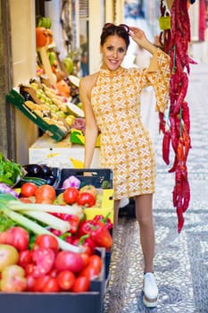Positive young female greengrocer in stylish short dress with hand touching eyeglasses on head while standing in town shop with fruit and vegetable on tray