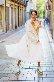 Smiling elegant young female wearing stylish coat and shoes standing on paved street against blurred old buildings and looking at camera