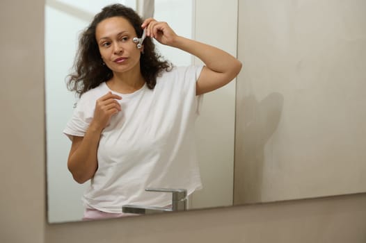 Attractive young multi ethnic woman with long curly brown hair, dressed in white pajama, looking at her mirror reflection while massaging her face with a facial roller massager, standing in bathroom