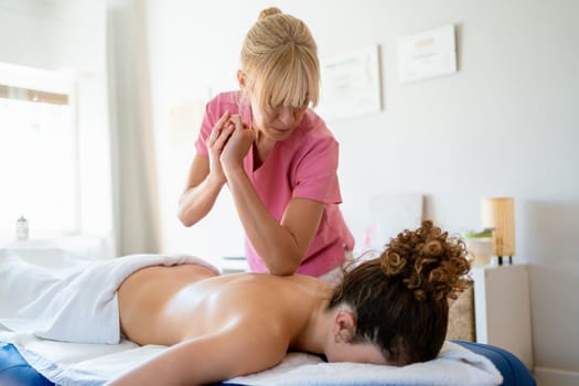 Concentrated female osteopath in uniform using elbow while massaging back of topless client lying face down on bed in modern physiotherapy clinic