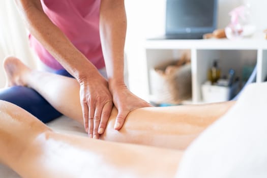 Crop female osteopathic therapist putting pressure and rubbing leg of female client lying on spa table during physiotherapist massage session in clinic