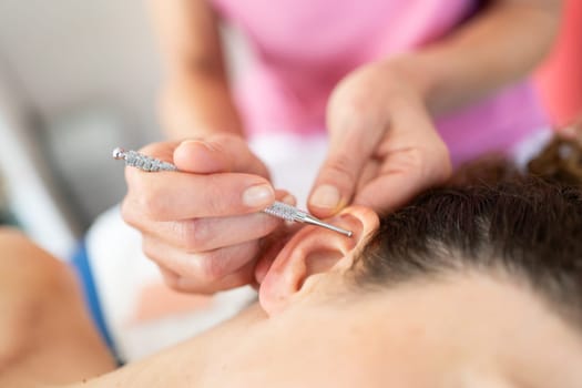 Crop professional cosmetician applying massage pen on ear of woman during auriculotherapy in modern beauty salon against blurred background