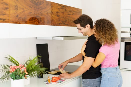 Side view of happy adult couple looking down while standing in kitchen near cooking range, smiling male in eyeglasses toasting bread slices on grill and female embracing from behind in daylight