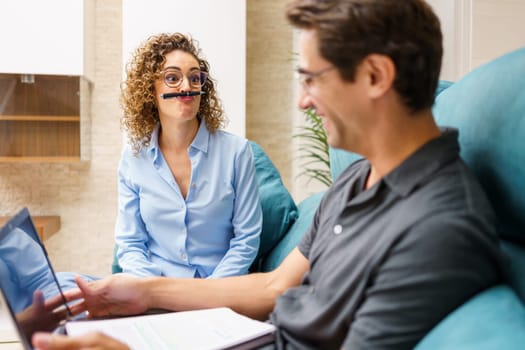 Positive young curly hair wife in smart casual shirt and eyeglasses playing with pen over mouth, while sitting on sofa and working with husband on remote project with laptop