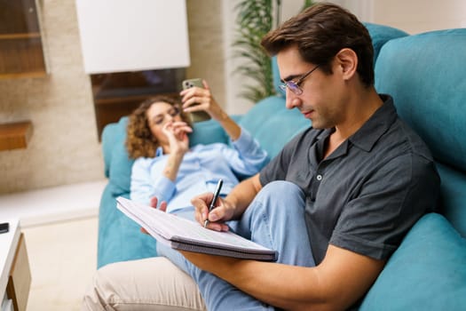 Side view of concentrated young man sitting on sofa and taking notes in notebook, while spending time with woman lying on couch with smartphone in living room
