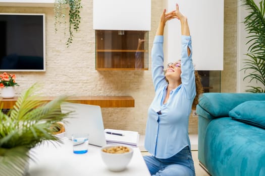 Happy young female freelancer in smart casual stretching raised arms while sitting near laptop in living room at home