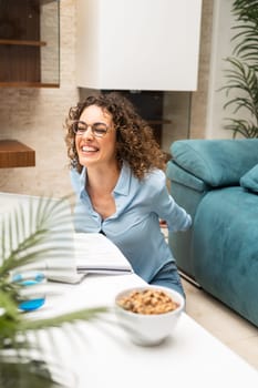 Cheerful young female freelancer in eyeglasses and smart casual working on project using laptop while sitting in living room at home