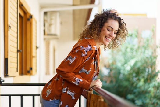 Side view of young curly haired happy female looking down while leaning on balcony railing at modern building against blurred background